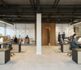 Modern open-plan office space with workstations, meeting rooms, and natural lighting from large windows. The office has a clean, industrial design with exposed ceilings, wooden accents, and a mix of seating options.