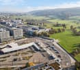 The image shows a panoramic view of a city with a mix of modern high-rise buildings and older structures. In the foreground, there is a large parking area with cars parked. The city is surrounded by rolling hills and countryside, creating a scenic contras