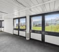 Large open office space with floor-to-ceiling windows offering a scenic view of the countryside outside. The room has a white ceiling with recessed lighting, gray carpeted floor, and black window frames. The space appears to be designed for a professional