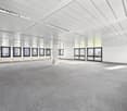 Large open office space with high ceilings, white walls, and gray carpeted flooring. The room has multiple windows along the walls providing natural light. There are several framed artworks or photographs hung on the walls.