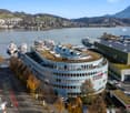 Circular blue building with green rooftop, located near waterfront with multiple ships and mountains in the background.
