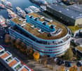 An office building with multiple floors, green rooftops, and solar panels on some parts. It is situated near the waterfront with boats docked nearby. The building is surrounded by various cars parked in parking lots. There are many other buildings and tre