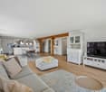living room with gray couch, white ottoman, wooden floor, white cabinets, TV, dining table, chairs, rug, and kitchen