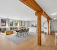 Dining room with wooden beams, large windows, dining table, chairs, rug, kitchen, wooden chest, white walls, wooden floor