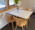 The image shows a dining room with a square wooden table and four wooden chairs. The table has a white marble-like top. There is a large window with curtains, providing natural light. A vase with a large green leaf and white flowers is placed on the table