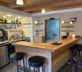 The image shows a cozy bar area with a wooden counter and shelves displaying various liquor bottles and glassware. The bar has a rustic, cabin-like feel with the wooden ceiling and walls. There is a coffee machine and a dishwasher visible, indicating this