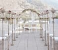 Outdoor wedding ceremony setup with a floral arch, white chairs, and a scenic mountain view in the background. The aisle is decorated with a patterned floor and candle holders lining the sides.