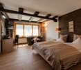 Spacious bedroom with wooden beams on the ceiling, wooden wall paneling, large windows, a seating area with a table and chairs, and a king-size bed with neutral-colored bedding.
