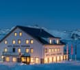 A large, multi-story hotel building with a balcony and terrace, located in a snowy, mountainous landscape with a clear blue sky. The hotel is illuminated at night, with several lit windows visible.