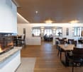 Spacious dining area with wooden floors, arched windows, and a fireplace. Tables and chairs are arranged throughout the room, and there are people seated at some of the tables. The lighting is warm and inviting, with drum-shaped light fixtures hanging fro