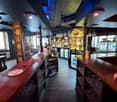A bar interior, with red countertops, shelves with glasses and bottles, wooden furniture, illuminated ceiling lights, and a blue-colored ceiling.
