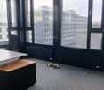 Large windows with a view of the surrounding buildings, gray carpeted floor, black framed windows, and a desk in the corner.