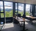 Large office space with floor-to-ceiling windows offering a scenic view of the surrounding landscape. The room features a long wooden desk with multiple computer monitors, chairs, and other office equipment. The space appears to be well-lit and modern in