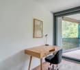 This image shows a modern and minimalist room with a wooden desk, a black chair, and a framed artwork on the wall. The room has large windows that provide a view of a balcony or terrace overlooking a wooded area.