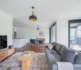 Open-concept living room with a large gray sofa, wooden coffee table, and a dining area with a wooden table and chairs. The room has a balcony or terrace with sliding glass doors, providing natural light and access to the outdoor space. The room features