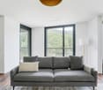 Spacious living room with large windows overlooking a forested area, hardwood floors, gray sofa, and a pendant light fixture.