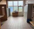 This image shows a room with a large window overlooking a scenic outdoor view. The room has tile flooring and contains various furniture and appliances, including a refrigerator, cabinets, and a wooden crate used for storage.