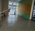 A large lobby with concrete flooring, a metal handrail on the stairs, a wall with green and brown paint, and glass doors leading to the outside with bicycles parked.