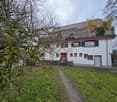 White building with red accents, wooden roof, balcony, garage, surrounded by a lawn and trees.