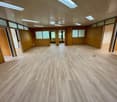 The image shows a large, open office space with a wooden floor and walls. The room has multiple windows and lighting fixtures, and appears to be a commercial or industrial space that could be used for various purposes such as an office, meeting room, or e