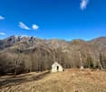 The image shows a picturesque mountain landscape with snow-capped peaks in the background. In the foreground, there is a small, rustic stone building surrounded by a bare, grassy field and a dirt path. The scene is framed by a forest of bare trees on eith
