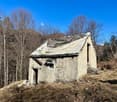 An old, dilapidated stone cabin with a slanted, slate roof in a forested, mountainous area. The cabin appears to be abandoned, with a small garage or outbuilding visible next to it.
