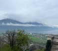 The image shows a scenic view of a mountainous landscape with clouds covering the peaks. There are trees in the foreground and a town or city visible in the valley below. The overall scene appears to be a picturesque view from a high vantage point, with t