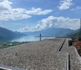 The image shows a scenic view of a lake surrounded by mountains. The foreground features a large gravel or pebble-covered roof or terrace, with a chimney or tower structure in the center. The lake and mountains in the background are visible through the cl