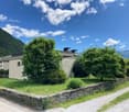 The image shows a scenic outdoor landscape with a grassy area, lush green trees and shrubs, and a stone wall surrounding the property. The sky is blue with fluffy white clouds, creating a picturesque natural setting.