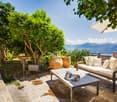 Lush green trees and foliage, stone patio with wooden furniture, large outdoor umbrella, mountain view in the background