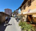 Narrow street with multistory buildings on both sides, a cyclist with a backpack, outdoor tables and chairs under awnings, potted plants, bicycles parked in front of the building, outdoor menu boards, parked truck on the side of the road.
