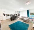 Modern living room with a blue rug, sofa, dining table, kitchen with appliances, and decorative items.