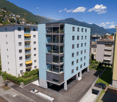 Multi-story apartment building with balconies, located in a mountainous area with scenic views. The building has a modern, minimalist design with a mix of white and gray colors.