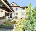 Traditional house with exposed stone walls, large wooden awning, wooden shelves, garden with various plants and trees, stone flooring