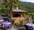 Outdoor dining area with a covered gazebo, palm trees, and tables set with purple tablecloths. There is a stone fireplace in the gazebo and the area is surrounded by lush greenery and mountains in the background.