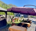 Outdoor dining area with a large purple umbrella, wooden tables and chairs, a small playhouse, and a scenic mountain backdrop.