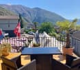 Terrace with table and chairs, fencing, potted plants, flags, view of mountains and houses.