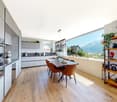 modern kitchen with wooden floor, large windows, dining table, wooden chairs, shelves with plants and books