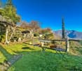 A garden with a table and chairs, surrounded by grass and plants, with a stone wall and a view of the mountains.