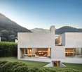 Modern two-story house with a minimalist concrete exterior, large windows, and a balcony overlooking a lush, mountainous landscape. The house also features a garage and offers a scenic view of the surrounding environment.