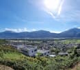 The image shows a panoramic view of a town or city surrounded by mountains. The town has a mix of residential and commercial buildings, with a modern-looking building visible in the foreground. The landscape is lush and green, with trees and vegetation co