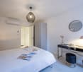 White bed with white sheets, 2 bedside lamps, air conditioner, round mirror, desk with hat and lamp, small table with trash can, cabinet, flower vase, flowers on bed, white tile flooring