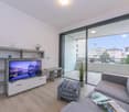 open living room with tv on concrete stand, gray sofa, ottoman, sliding doors to balcony, glass windows, hardwood floor, plants, lamp, cityscape view