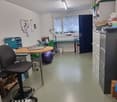 The image shows a small office or workspace. It has a desk with a computer, a printer, and other office equipment. There is also a washing machine visible, indicating that this space may be used for both work and personal activities.