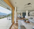 modern living room with large windows, wooden floor, gray couch, wooden coffee table, round table with chairs, kitchen area, balcony with table and chairs, view of the city and mountains