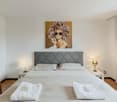 Spacious bedroom with a large bed, gray upholstered headboard, and a painting of a woman with a floral hairstyle on the wall. The room also features wooden floors, bedside tables with lamps, and a clothing rack.