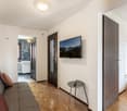 Open-plan studio apartment with hardwood floors, white walls, large windows, sofa, coffee table, TV mounted on the wall, bathroom with tiled walls, and a kitchenette area with a dishwasher.