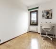 Spacious bedroom with hardwood flooring, white walls, a large window, and a desk with a chair. The room also features shelving and a decorative artwork on the wall.