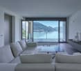 open living space, large grey sofa, glass coffee table, large glass windows, balcony, lake and mountain view