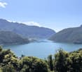 The image shows a scenic view of a lake surrounded by mountains and lush greenery. The lake appears to be a deep blue-green color, reflecting the sky and the surrounding landscape. The mountains in the background are covered in dense vegetation, creating